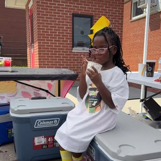 Brielle, the young founder, in a yellow bow and pink glasses, enjoying a treat at her lemonade stand