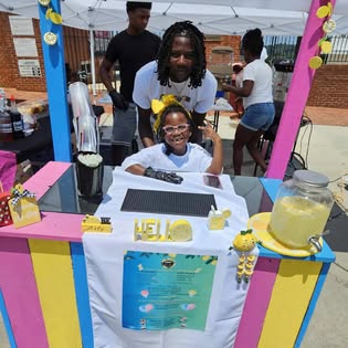 Brielle at her decorated BKrazy stand under a tent, with family and jars of fresh lemonade